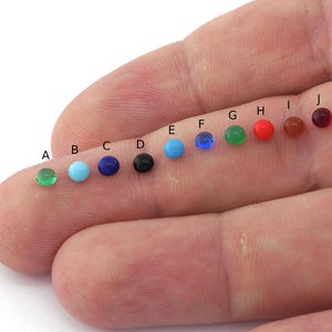 Könnte beinhalten: Eine Reihe von zehn kleinen runden Perlen in verschiedenen Farben, darunter Grün, Türkis, Blau, Schwarz, Rot und Braun. Die Perlen sind in einer Linie auf einem Finger angeordnet.
