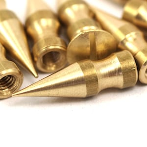 May include: Close-up of a group of brass-colored, pointed, threaded metal tips. The tips are arranged in a random pattern on a white surface.