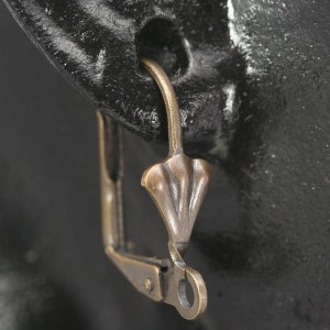 May include: A gold-toned earring with a unique, intricate design. The earring features a delicate, floral-shaped pendant with a curved, open-ended wire.