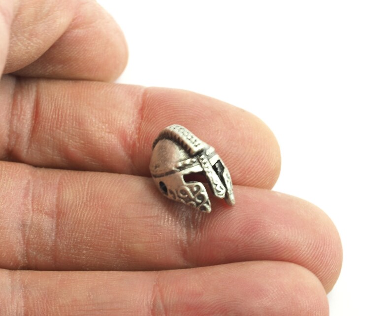 May include: A silver-colored metal charm in the shape of a helmet. The helmet has a detailed design and is a popular choice for jewelry making.