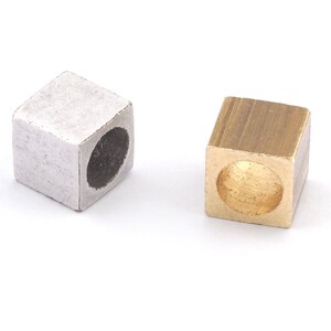 Op de afbeelding: Twee kubusvormige metalen kralen. Eén is zilverkleurig, de andere goudkleurig. Elke kraal heeft een groot, rond gat in het midden. Deze kralen zijn geschikt voor het maken van sieraden en knutselen.