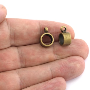 Op de afbeelding: Twee antieke hangerbeugels in messingkleur. Elke beugel heeft een cirkelvormige ring met een kleine lus aan de bovenkant om aan een ketting of een ander sieradenonderdeel te bevestigen. De beugels worden tegen een lichte achtergrond getoond.