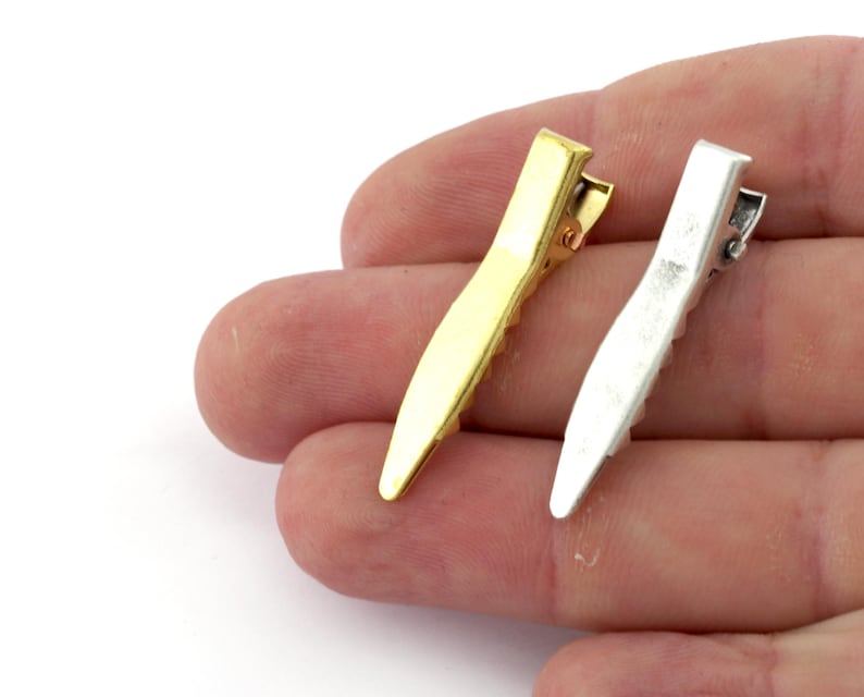 K&ouml;nnte beinhalten: Zwei Haarklammern, eine goldene und eine silberne, mit einer gezahnten Kante. Die Klammern werden in einer Hand gehalten.
