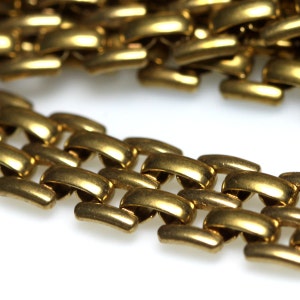 Puede incluir: Primer plano de un collar de cadena dorado. Los eslabones de la cadena son de forma ovalada y están entrelazados, creando una superficie texturizada. El collar está sobre un fondo blanco, resaltando el brillo metálico.