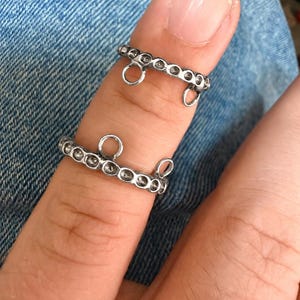 May include: Two silver-toned rings featuring a series of circular indentations along the band. Each ring includes two small, circular loops extending from the top edge, designed for attaching additional jewelry components or charms.