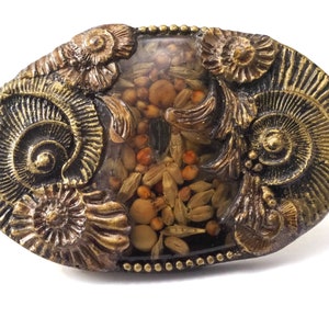 May include: An oval-shaped, bronze-colored resin object with embedded seeds and small stones. The edges are adorned with detailed, spiraling fossil designs. The center is filled with a clear resin, showcasing the natural elements within.