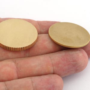 Op de afbeelding: Twee ronde, goudkleurige metalen schijven. De randen van de schijven hebben een geribbelde textuur, terwijl de vlakken glad zijn. De schijven worden op een lichtgekleurde achtergrond getoond.