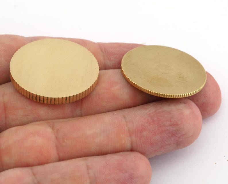 Op de afbeelding: Twee ronde, goudkleurige metalen schijven. De randen van de schijven hebben een geribbelde textuur, terwijl de vlakken glad zijn. De schijven worden op een lichtgekleurde achtergrond getoond.