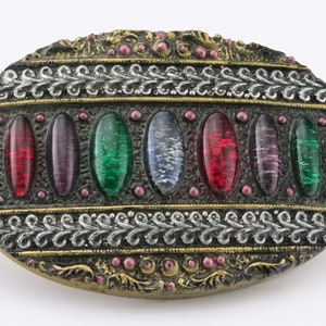 May include: An oval-shaped, antique-style brooch with a dark metallic frame. The brooch features a row of colorful, oval-shaped glass stones in shades of blue, red, purple, and green. The frame is decorated with silver and gold-colored filigree and small pink accents.