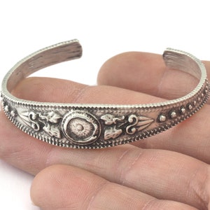 Könnte beinhalten: Ein silbernes Armband mit einem verzierten Design, das einen zentralen Kreis und florale Motive aufweist. Das Armband ist offen und hat eine einfache, abgerundete Form.