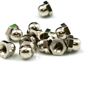 Puede incluir: Un conjunto de tuercas ciegas plateadas brillantes, también conocidas como tuercas de campana, se dispersan sobre un fondo blanco. Estas tuercas hexagonales tienen una parte superior redondeada en forma de cúpula y un interior roscado para una fijación segura.