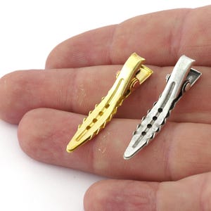 K&ouml;nnte beinhalten: Zwei Haarklammern, eine goldfarben und eine silberfarben, mit einem strukturierten Design. Die Klammern werden in einer Hand gehalten.