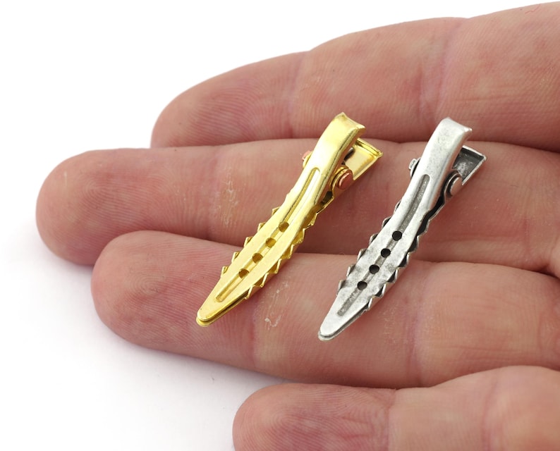 K&ouml;nnte beinhalten: Zwei Haarklammern, eine goldfarben und eine silberfarben, mit einem strukturierten Design. Die Klammern werden in einer Hand gehalten.