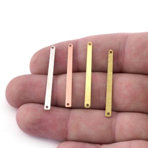 May include: Four metal bar charms in silver, copper, gold, and textured brass. Each bar has a hole at both ends, ideal for jewellery making. The bars are held in a hand against a white background.