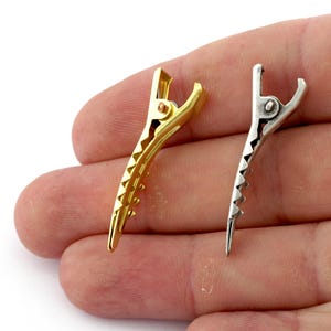 K&ouml;nnte beinhalten: Zwei Haarklammern, eine goldene und eine silberne, mit einem gezahnten Griff. Die Klammern werden in einer Hand gezeigt.
