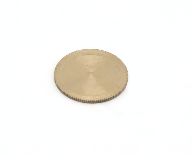 Op de afbeelding: Een rond, messingkleurig metalen object met een geribbelde rand. Het object is cirkelvormig en heeft een licht getextureerd oppervlak. De achtergrond is effen wit.