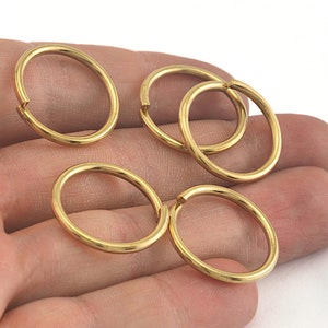 Puede incluir: Se muestran varios anillos de salto ovalados de color dorado. Los anillos están abiertos, con un pequeño espacio en un punto del círculo. Los anillos son lisos y brillantes, reflejando la luz. El fondo es una superficie blanca lisa.