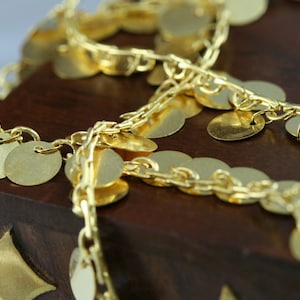May include: Gold chain with round, flat, coin-like charms. The chain is on a dark brown wooden surface.