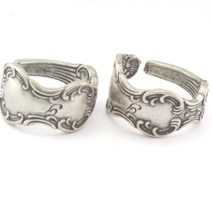 Puede incluir: Dos anillos plateados hechos de cubiertos ornamentados. Cada anillo presenta una cara rectangular decorativa con adornos de volutas. Los anillos se muestran sobre un fondo blanco, resaltando sus intrincados detalles y su estética vintage.
