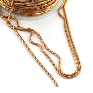Könnte beinhalten: Ein durchgehendes Stück goldfarbene Schlangenkette, auf eine weiße Spule gewickelt. Die Kette zeichnet sich durch ein glattes, flexibles und abgerundetes ineinandergreifendes Glieddesign mit metallischem Finish aus, das in losen Strängen abfällt.