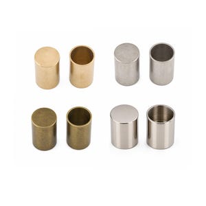 May include: A collection of eight cylindrical metal caps arranged in two rows. The set includes pairs with smooth finishes in polished gold, brushed silver, antique bronze, and a reflective chrome-like silver, each featuring a hollow interior.