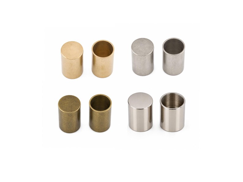 May include: A collection of eight cylindrical metal caps arranged in two rows. The set includes pairs with smooth finishes in polished gold, brushed silver, antique bronze, and a reflective chrome-like silver, each featuring a hollow interior.