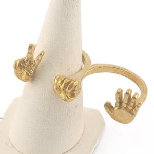May include: Gold ring with two hands, one making a peace sign and the other an open hand, on a white display stand.