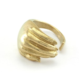 May include: A gold-colored ring in the shape of a hand, with detailed finger ridges. The ring is open at the back, allowing for size adjustment. The ring is set against a white background.
