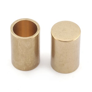 Pode incluir: Duas buchas cilíndricas de bronze, uma oca com rosca interna, a outra maciça, sobre fundo branco. Possuem um acabamento metálico liso, provavelmente para máquinas ou aplicações mecânicas. Cada bucha tem cerca de 2,5 cm de altura.