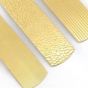 Peut inclure: Trois bandes métalliques rectangulaires de couleur dorée disposées en diagonale. La bande de gauche présente une texture finement quadrillée, celle du milieu un motif martelé en relief, et celle de droite des lignes diagonales parallèles.