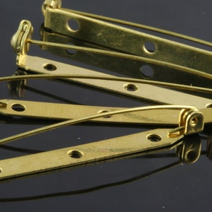 Pode incluir: Um conjunto de quatro costas de broche de metal dourado com fecho de pino de segurança. As costas de broche estão dispostas em uma pilha sobre uma superfície preta.