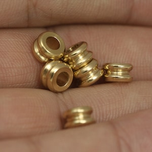 May include: A close-up of five gold-coloured metal beads. The beads are round and have a smooth, polished finish. They are arranged in a pile on a person's hand.