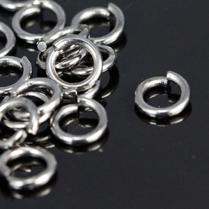 May include: A close-up of a pile of silver jump rings. The rings are small and round, and they are all linked together. The rings are on a black surface.
