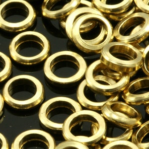 Puede incluir: Primer plano de numerosos anillos metálicos pequeños de color latón. Los anillos son circulares con un agujero en el centro y un borde exterior ligeramente elevado. Los anillos están dispersos sobre una superficie oscura.