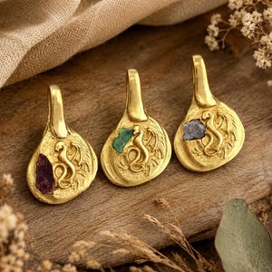 May include: Three gold-toned metal pendants, each featuring a raised dragon relief and an embedded raw gemstone. The first pendant contains a dark red stone, the second a green stone, and the third a dark grey stone. Each piece has a smooth bail.