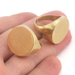 May include: Two gold-colored signet rings with a brushed finish. The rings have a flat, oval face and a simple band. The rings are displayed on a white background.