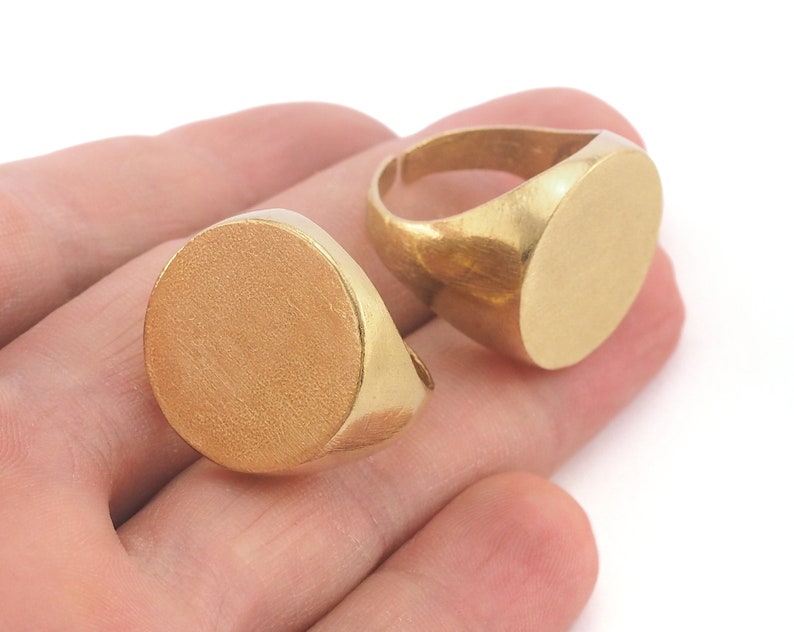 May include: Two gold-colored signet rings with a brushed finish. The rings have a flat, oval face and a simple band. The rings are displayed on a white background.