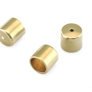 May include: Three small, cylindrical brass objects with a polished finish are arranged on a white surface. Each cylinder has a flat top and bottom, with a small hole in the top. One cylinder has an open end.