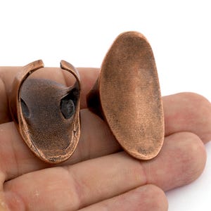 May include: Two copper rings, one with a smooth oval shape and the other with a more intricate design. Both rings are shown on a hand.