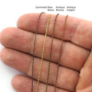 Puede incluir: Primer plano de cuatro cadenas de bolas en colores gunmetal, latón crudo, bronce antiguo y cobre antiguo. Las cadenas se muestran sobre una mano, mostrando los diferentes acabados metálicos. El texto sobre las cadenas dice "Gunmetal Raw Brass Antique Bronze Antique Copper".