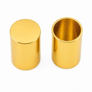 May include: Two cylindrical metal caps with a polished gold finish. One cap is shown from the top, displaying a solid flat circular surface, while the other is positioned to reveal its hollow interior with visible internal threading along the wall.