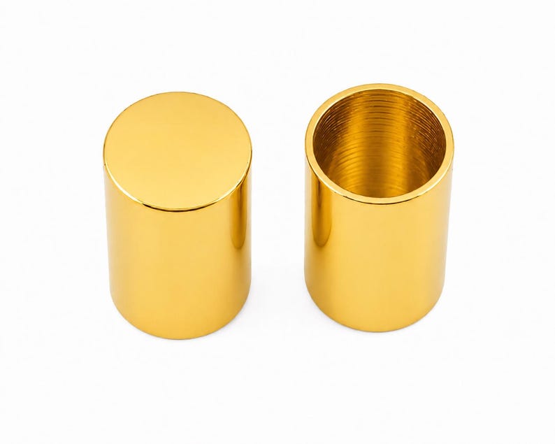 May include: Two cylindrical metal caps with a polished gold finish. One cap is shown from the top, displaying a solid flat circular surface, while the other is positioned to reveal its hollow interior with visible internal threading along the wall.