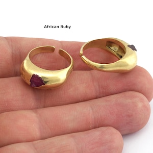 Puede incluir: Dos anillos dorados con una gema de rubí en bruto incrustada. Los anillos tienen un diseño redondeado y ajustable. El texto "African Ruby" está encima de los anillos. Se muestran en una mano.