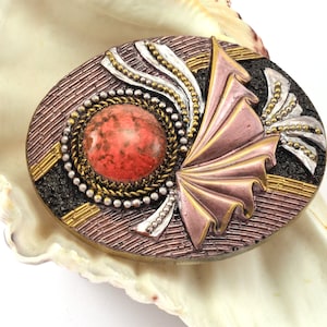 May include: An oval-shaped brooch with a pink, gold, and black design. The center of the brooch features a large, round, red stone.