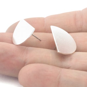 Pode incluir: Um par de brincos de botão prateados. Cada brinco tem um design semicircular liso com uma borda plana e um pino para perfurar. Os brincos são exibidos em um fundo neutro.