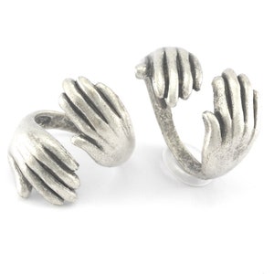 May include: Two silver rings shaped like hands, each hand is holding the other's wrist, creating a heart shape.