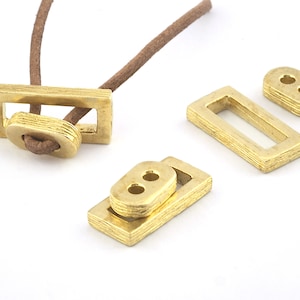 Puede incluir: Cuatro cierres rectangulares de metal dorado con dos agujeros. Dos cierres están apilados uno encima del otro. Un cierre está enhebrado con un cordón marrón.