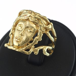 Puede incluir: Un anillo de oro con un diseño detallado de la cara de una mujer rodeada de un patrón texturizado e intrincado.