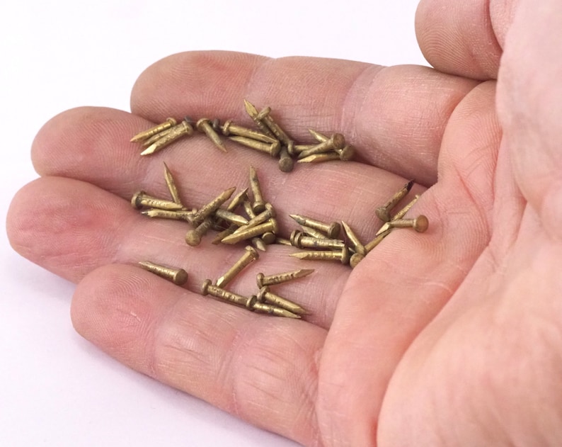 May include: A hand holding a pile of small, gold-colored tacks or nails.