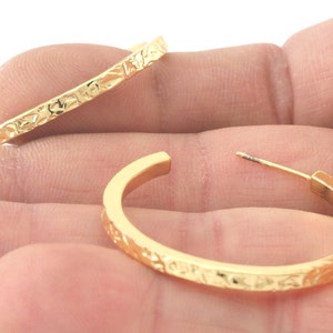 May include: A pair of gold hoop earrings with a textured surface. The earrings are shown on a hand.